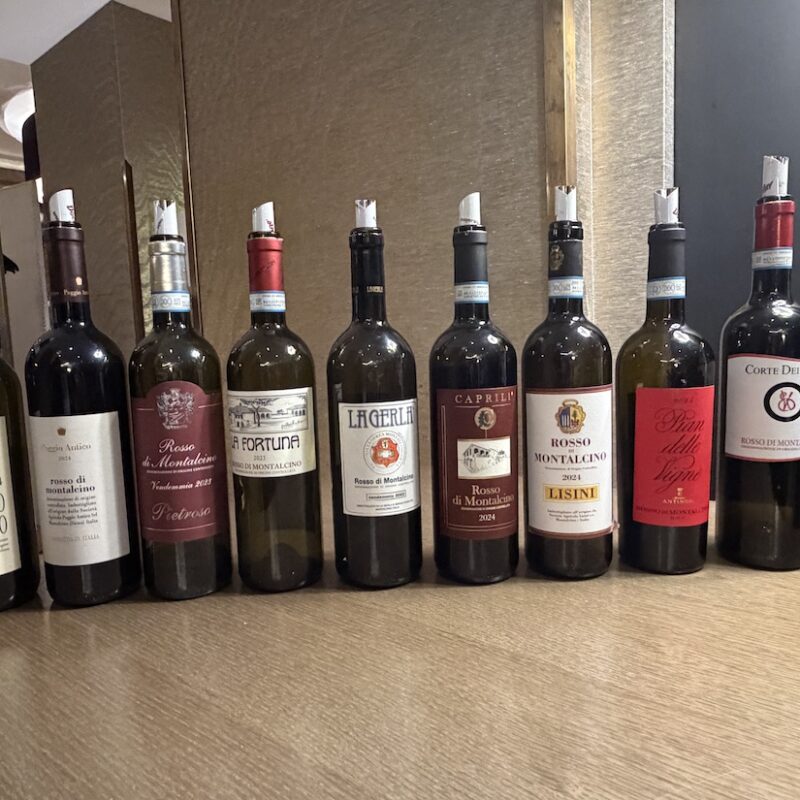 Allineamento di bottiglie di vino Rosso di Montalcino di diversi produttori sul tavolo della degustazione DoctorWine.