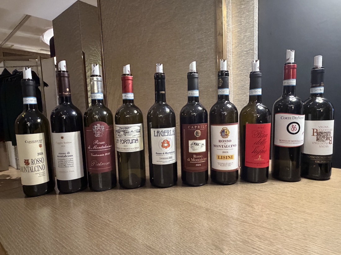 Allineamento di bottiglie di vino Rosso di Montalcino di diversi produttori sul tavolo della degustazione DoctorWine.